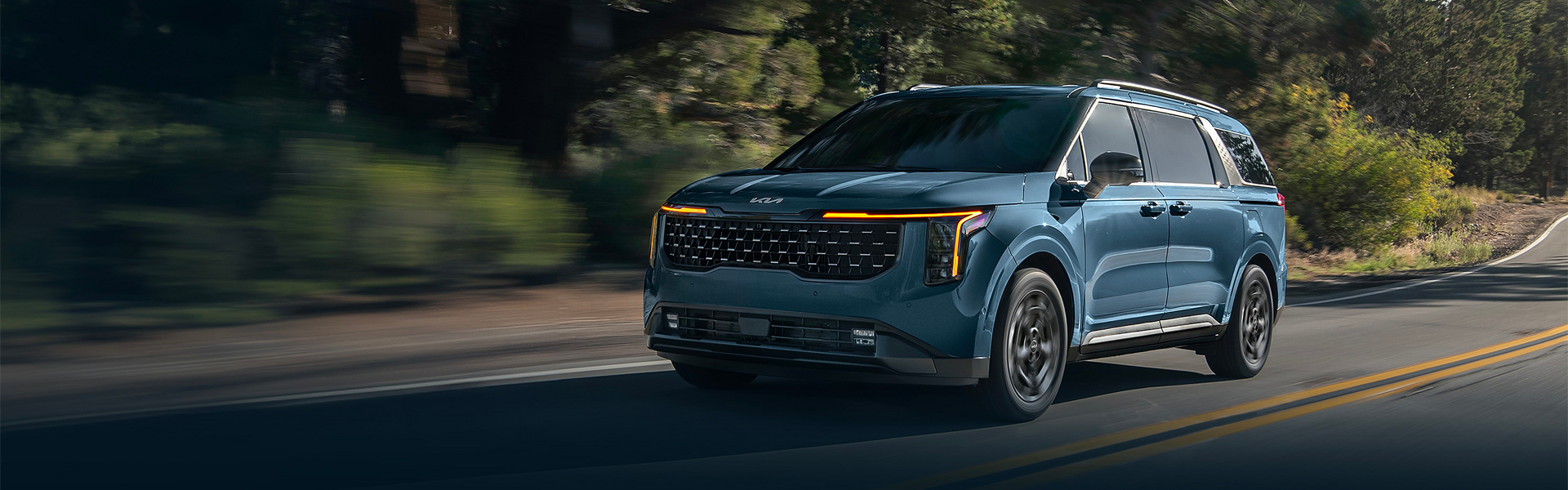 A blue Kia Carnival minivan driving on a winding, tree-lined road with bright headlights.