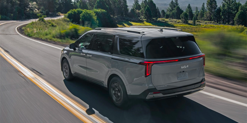 Gray Kia Carnival minivan with roof rails driving on a winding road with trees in the background.