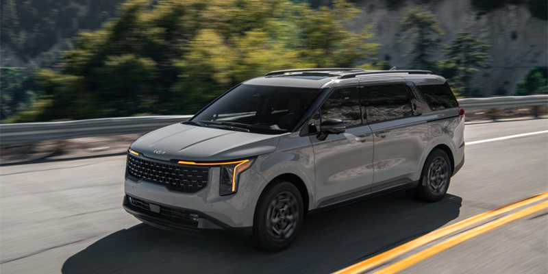 A light gray Kia Carnival minivan drives right on a paved road with blurred green trees and blue sky in the background.