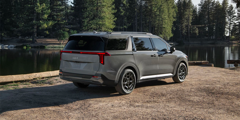 A gray Kia Carnival minivan is parked on a dirt path next to a serene lake, surrounded by towering pine trees.