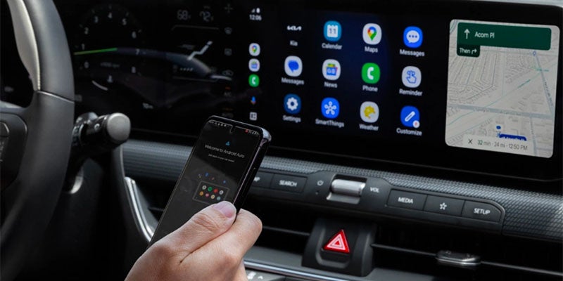 A hand holds a smartphone connecting to a car's infotainment screen, displaying app icons and a map.