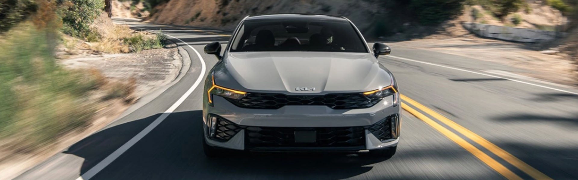 A silver Kia sedan drives toward the viewer on a winding road with a muted mountainous background.