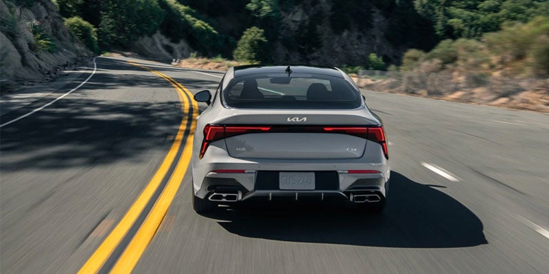 Rear view of a grey Kia K5 driving on a winding road with trees and hills in the background.