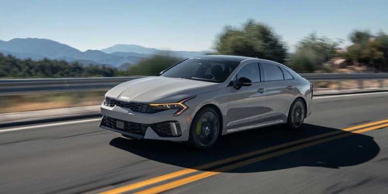 Light gray car with headlights on, driving on a highway with mountains in the background.