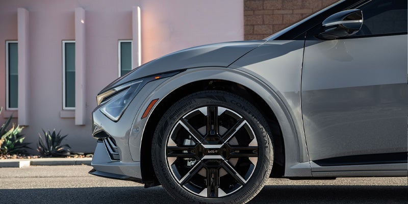 Front side of a gray car parked on an asphalt street with a light pink building and plants in the background.