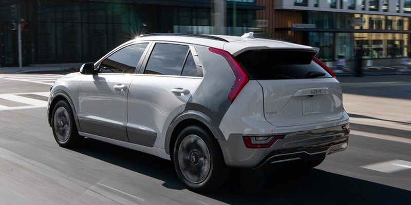 A white Kia Niro EV drives right on a city street. The rear and right side of the car are visible.