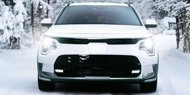 Front view of a white Kia car with bright headlights on a snowy road with snow-covered trees in the background.