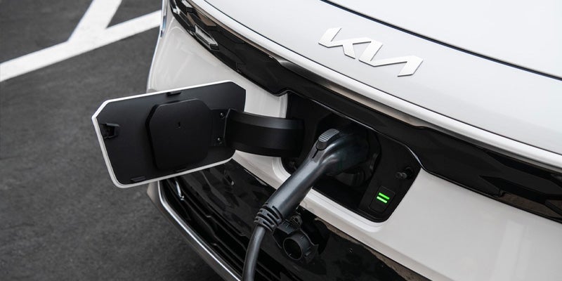 A white Kia EV being charged with a black charging cable. The charge port door is open.