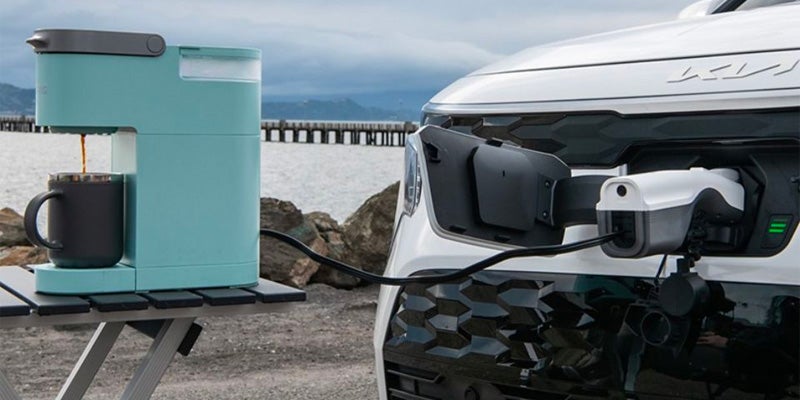 A teal coffee maker brewing coffee into a mug, powered by an electric vehicle charger connected to a white car. Waterfront background.