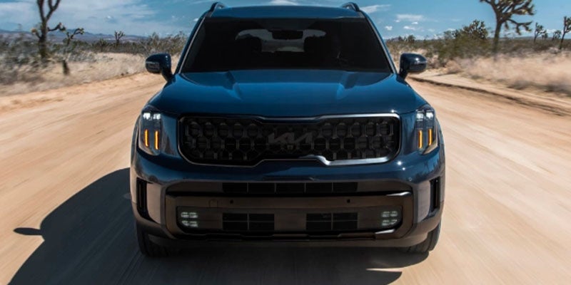 Front view of a dark blue SUV with black grille and illuminated vertical daytime running lights, driving down a dirt road.