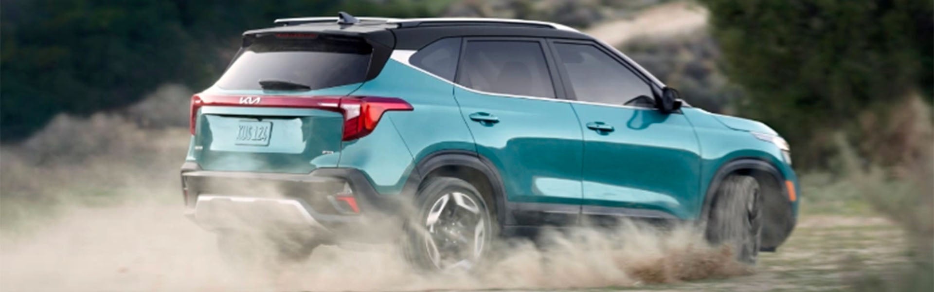 Rear view of a teal SUV driving on a dirt road, kicking up dust. The roof is black and the taillights are red.