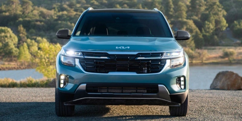 Front view of a teal Kia SUV with its headlights on, parked on a gravel road next to a body of water.