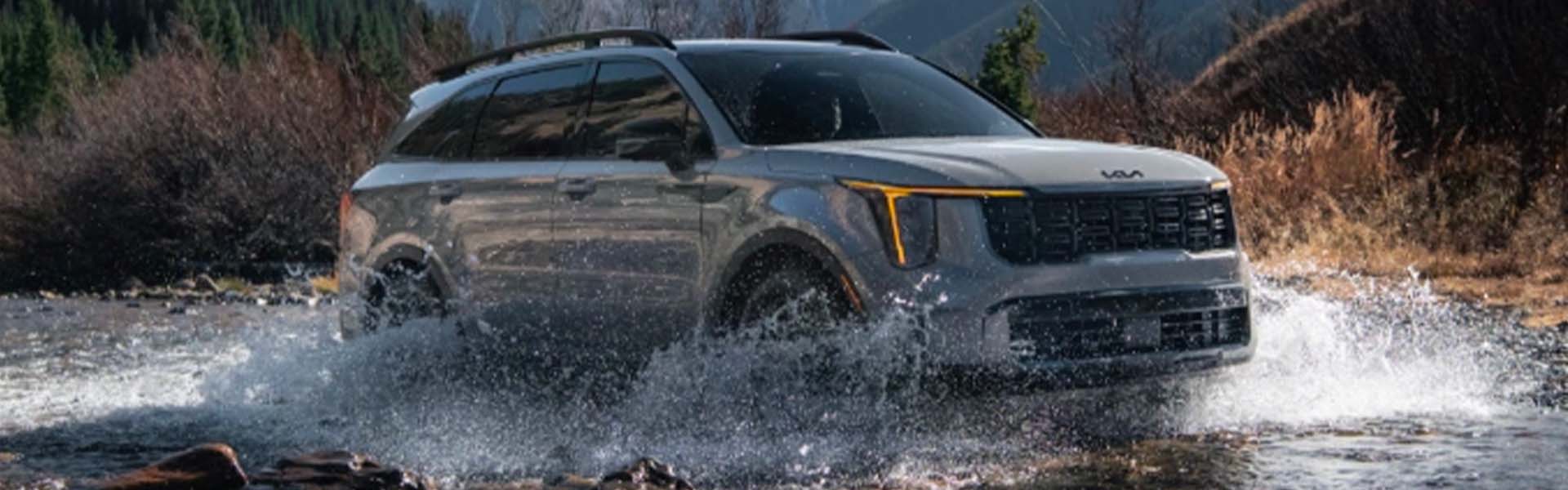 A gray SUV with yellow daytime running lights driving through a river, splashing water. Mountains and trees are in the background.
