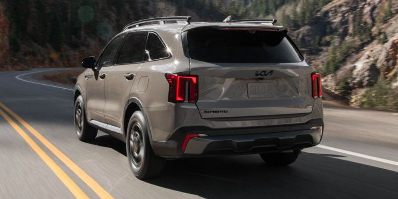Rear view of a light gray Kia Sorento driving on a winding road with mountains and trees in the background.