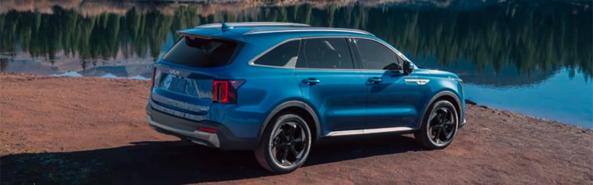 A blue SUV parked sideways on a dirt path next to a serene lake reflecting trees.