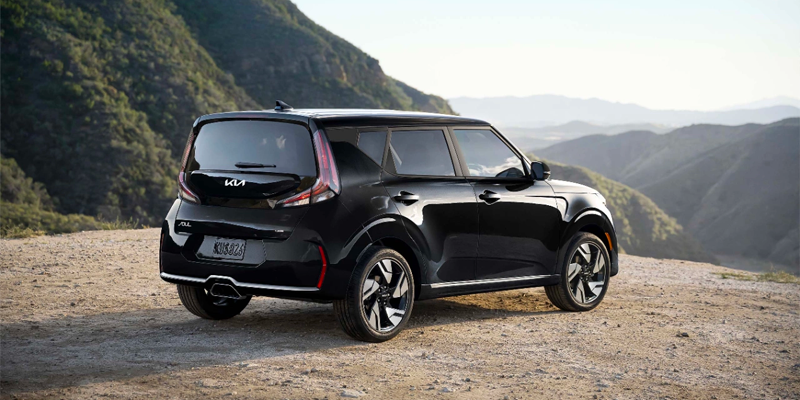 A black Kia Soul parked on a dirt road, viewed from the rear passenger side, with hills and mountains in the background.