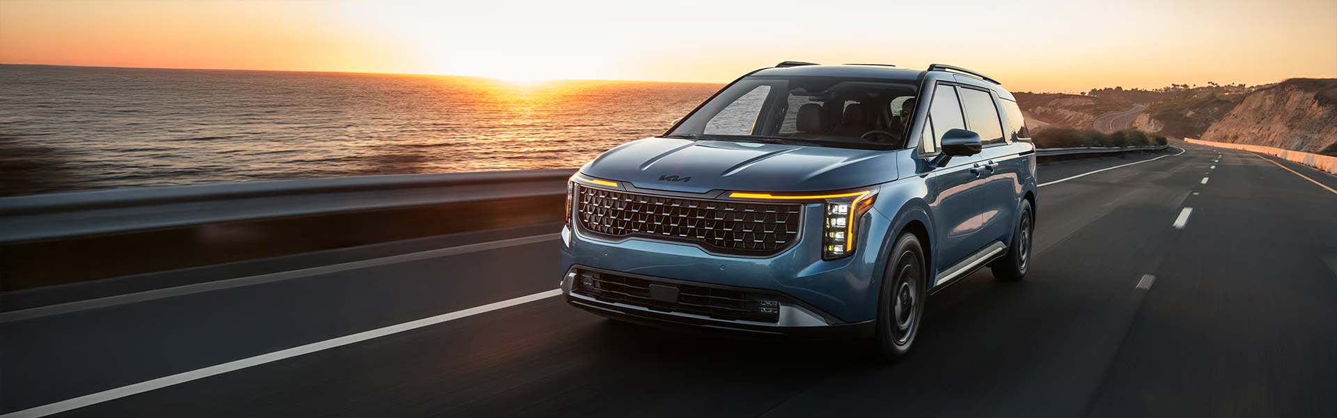 A blue Kia Carnival minivan drives forward on a highway next to the ocean at sunset.