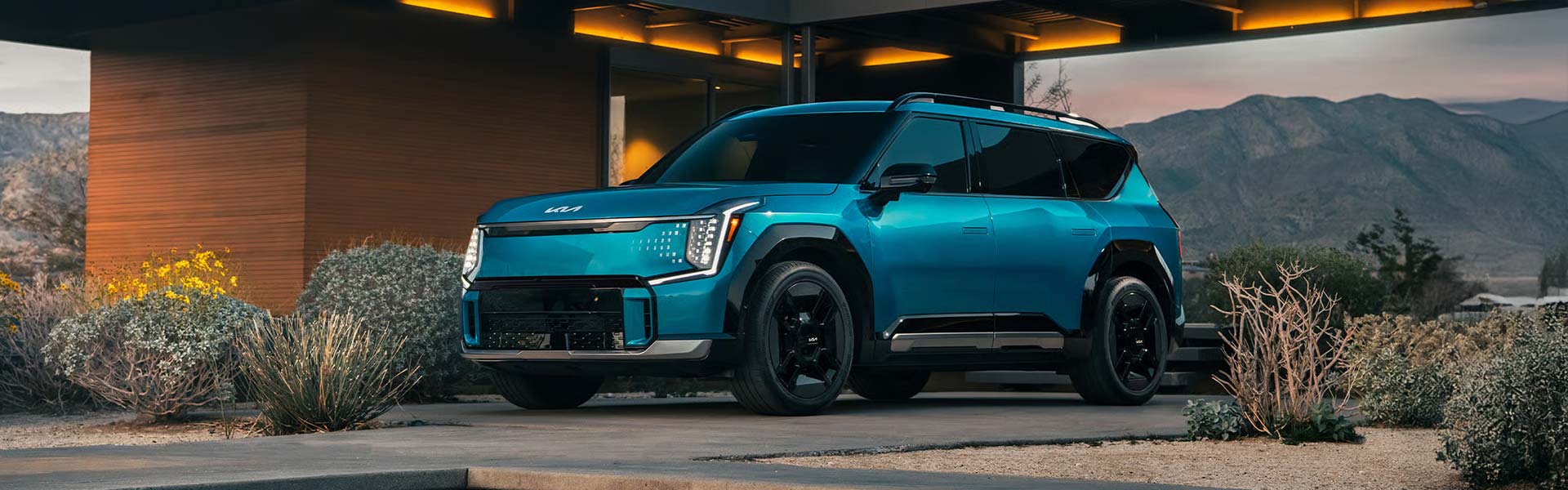 Kia EV9 parked beneath a modern wooden home with mountains in the background.