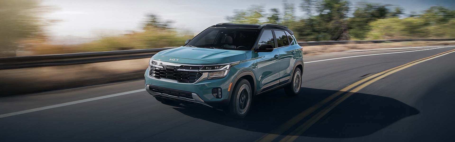 A teal Kia Seltos drives on a highway, with blurred green trees in the background.